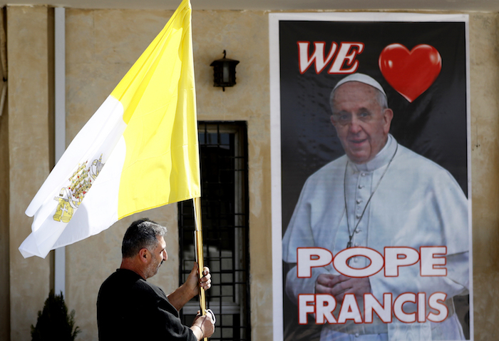 Un sacerdote cristiano sostiene una bandera del vaticano mientras camina ante un cartel del Papa Francisco, durante los preparativos para la visita del papa en Mar Youssif Church en Bagdad, Irak, el viernes 26 de febrero de 2021. 