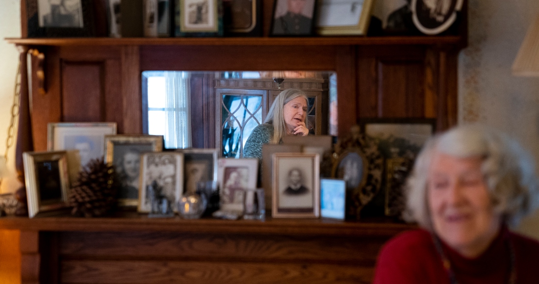 Nancy Rose, en el centro en el espejo, habla con su madre, Amy Russell, a la derecha, en su comedor rodeadas de fotografías de amigos familiares, el martes 25 de enero de 2022 en Port Jefferson, Nueva York. Ambas contrajeron COVID-19.