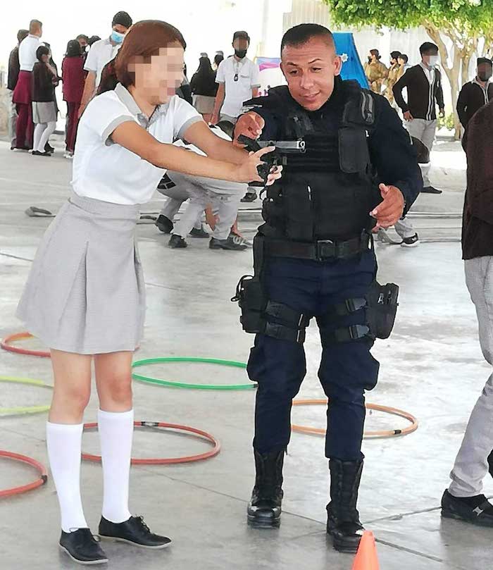 Una Adolescente Empuña Una Pistola Rodeada De Compañeras Y Compañeros Mientras Un Policía Uniformado Levanta El Brazo Y Apunta Por Arriba Del Arma Aparentemente Haciendo Alguna Indicación a La Persona Menor De Edad