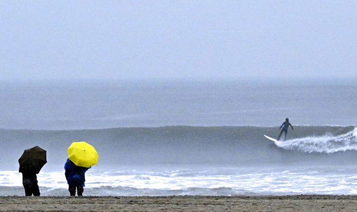 Unas personas caminan junto al mar mientras surfistas montan las olas durante una tormenta en Venice Beach, en Los Ángeles, California, el sábado 14 de enero de 2023. 