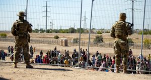 Migrantes esperan junto a una valla fronteriza mientras son vigilados por miembros de la Guardia Nacional de Texas, para ingresar a El Paso, Texas, el miércoles 10 de mayo de 2023.