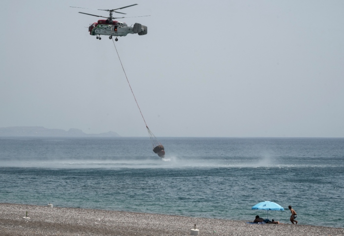 Dos personas en la playa observan como un helicóptero carga una cesta de agua en el mar en un operativo de extinción de incendios, cerca de Gennadi, en la isla de Rodas, en el sureste de Grecia, el 27 de julio de 2023. Foto: Petros Giannakouris, AP