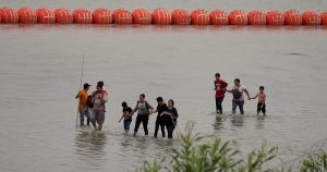 Migrantes que cruzaron el Río Grande (Bravo) desde México caminan frente a enormes boyas colocadas como una barrera fronteriza flotante en el río, el miércoles 12 de julio de 2023, en Eagles Pass, Texas.