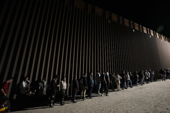 Varias personas se forman junto a un muro fronterizo mientras esperan solicitar asilo tras cruzar la frontera desde México, el martes 11 de julio de 2023, cerca de Yuma, Arizona.