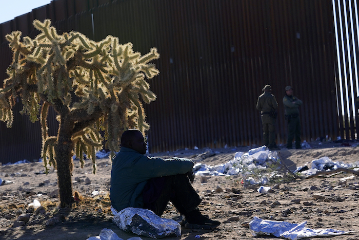 Un migrante se sienta a la sombra de un cactus saltarín del desierto mientras se une a cientos de migrantes reunidos a lo largo de la frontera, el martes 5 de diciembre de 2023 en Lukeville, Arizona.