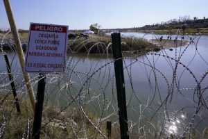 Migrantes cruzan el Río Bravo a Estados Unidos desde México detrás de un hilo de cuchillas y un cartel que advierte que cruzar es peligroso e irregular, el miércoles 3 de enero de 2024 en Eagle Pass, Texas.