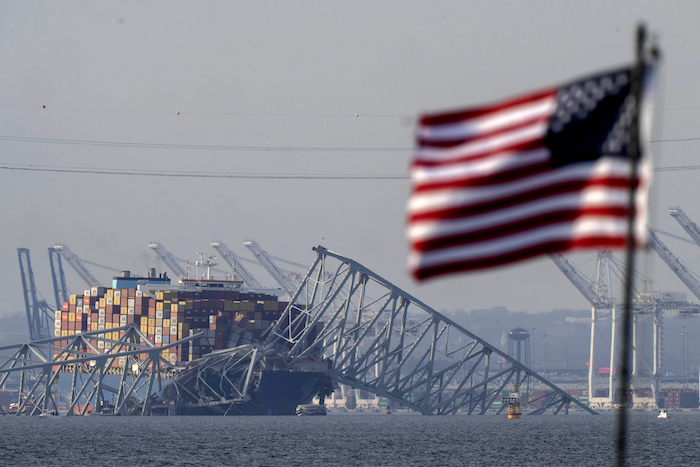 Una bandera estadounidense ondea en primer plano mientras, en el fondo, el portacontenedores Dalí descansa contra los restos del puente Francis Scott Key, el 26 de marzo de 2024, visto desde Pasadena, Maryland.