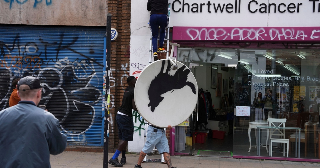 Personas retiran una nueva obra de arte de Banksy, que representa un lobo aullando pintado en una antena parabólica que se colocó en el techo de una tienda en Peckham, al sur de Londres, el jueves 8 de agosto de 2024. 